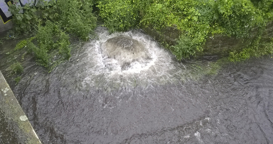 Wasser quillt aus einer Schachtabdeckung. Die Ursache ist üblicherweise Rückstau aus dem Kanal als Folge eines Starkregens. Bei unterirdischen Regenspeichern unterhalb der Rückstauebene, die nicht dagegen gesichert sind, droht die Kontamination des Wasservorrats.