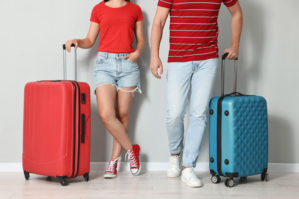 Zwei Menschen stehen mit Koffern da, leger gekleidet in roten Oberteilen und Jeanshosen, bereit zum Reisen. - © New Africa - stock.adobe.com