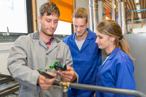 Drei SHK-Techniker in blauen und grauen Overalls begutachten ein Heizsystemgerät in einer Werkstattumgebung mit verschiedenen Rohrinstallationen zur Analyse der Energieeffizienz. - © Atelier 211 - stock.adobe.com