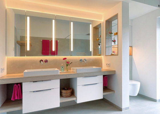 „Modernes Badezimmer mit zwei Waschbecken und hinterleuchteten Spiegeln, die energieeffiziente Beleuchtungstechniken veranschaulichen. Rosa Handtücher und dekorative Regale demonstrieren ästhetische Gestaltungsmöglichkeiten für nachhaltige Renovierungen im SHK-Bereich. - © Bild: Hansen