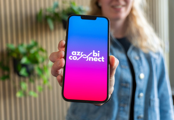 Eine Person hält ein Telefon, auf dem das Logo „azco bi connect“ auf einem blau-rosa Farbverlaufsbildschirm angezeigt wird. - © Kreishandwerkerschaft Oldenburg