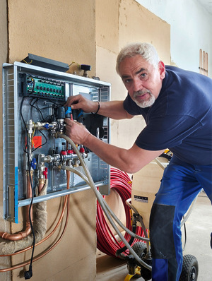 Ein Techniker mit grauem Haar arbeitet an Rohren und Ventilen in einem offenen Wandpaneel und blickt in die Kamera. - © Bild: Moritz Hartmann