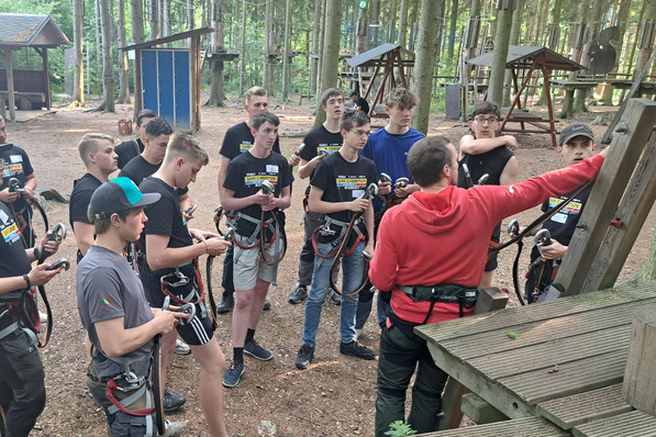 Eine Gruppe Jugendlicher in Klettergurten hört einem Ausbilder in einem roten Kapuzenpulli in einem Outdoor-Seilgarten zu. - © Fachverband SHK Sachsen