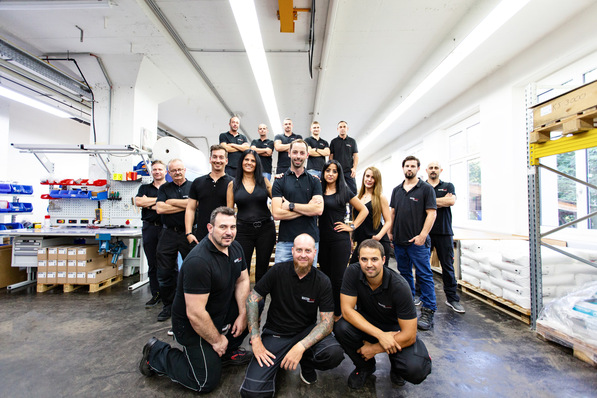 Eine Gruppe von Menschen in schwarzen Hemden posiert zusammen in einem hellen industriellen Arbeitsraum. - © Chris Reichl