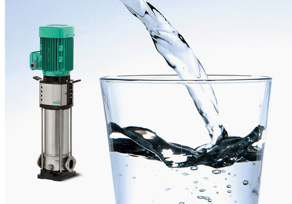 Ein Glas Wasser, das gefüllt wird, neben einer grünen und silbernen industriellen Wasserpumpe auf einem hellen Hintergrund. - © Bild: Wilo SE
