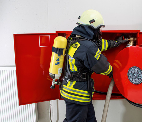Ein Feuerwehrmann in voller Montur benutzt einen Feuerwehrschlauch aus einem an der Wand montierten Schlauchkasten. - © Bild: Wilo SE