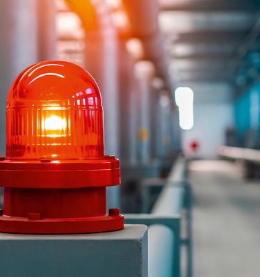 Eine rote Warnleuchte blinkt in einer Industrieanlage mit verschwommenen Rohren im Hintergrund. - © Bild: Yauhen - stock.adobe.com