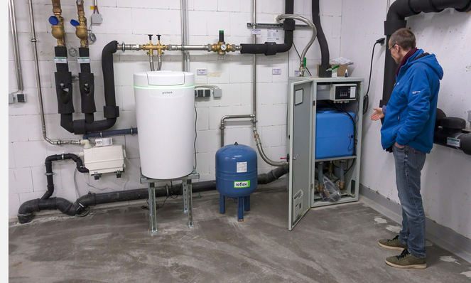 Ein Mann steht in einem Hauswirtschaftsraum mit Heizungs- und Wasserversorgungsanlagen und Rohren an den Wänden.