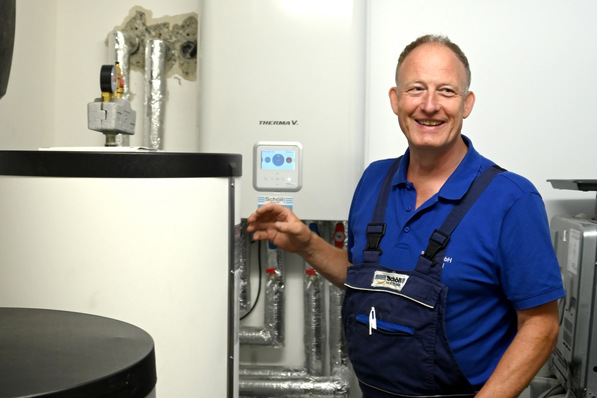 Ein lächelnder Techniker in blauer Uniform steht neben einer Heizungsanlage in einem Versorgungsraum. - © Reisser